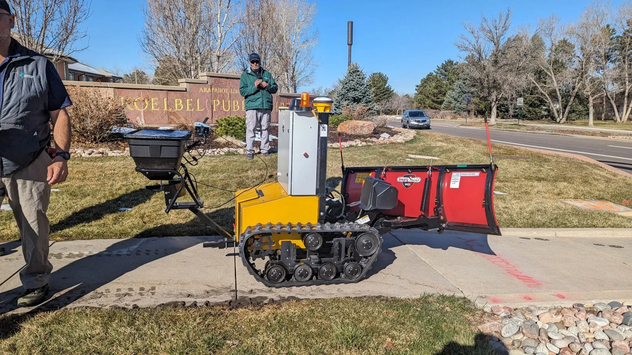 A Snowbotix SMUR unit on a paved library path during the Arapahoe Libraries pilot in Colorado, with two crew members alongside.