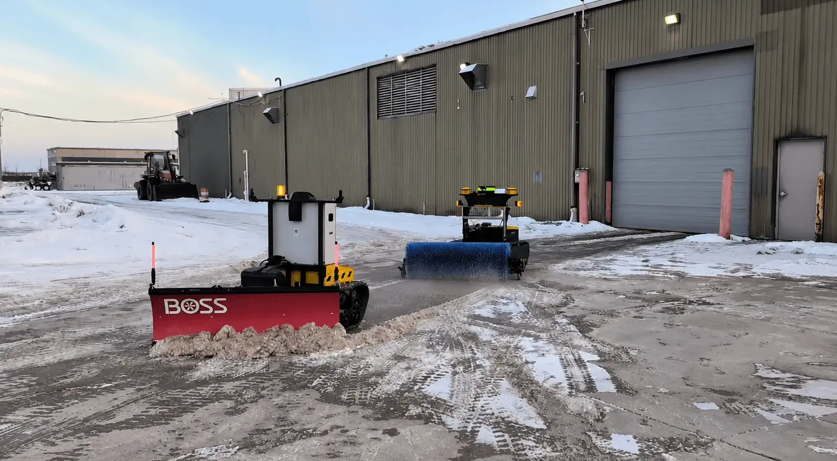 Two Snowbotix SMUR units with front plow blades clearing snow outside a commercial warehouse before opening.