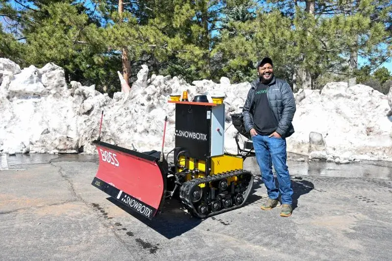 A Snowbotix SMUR unit with a front plow blade beside a crew member during the City of Greeley sidewalk pilot, with cleared snow piled behind.